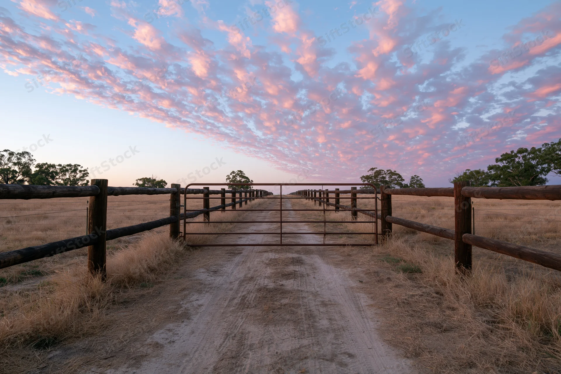 Metal farm gate flanked by dark timber post-and-rail fences on a dirt road leading into a field at sunset