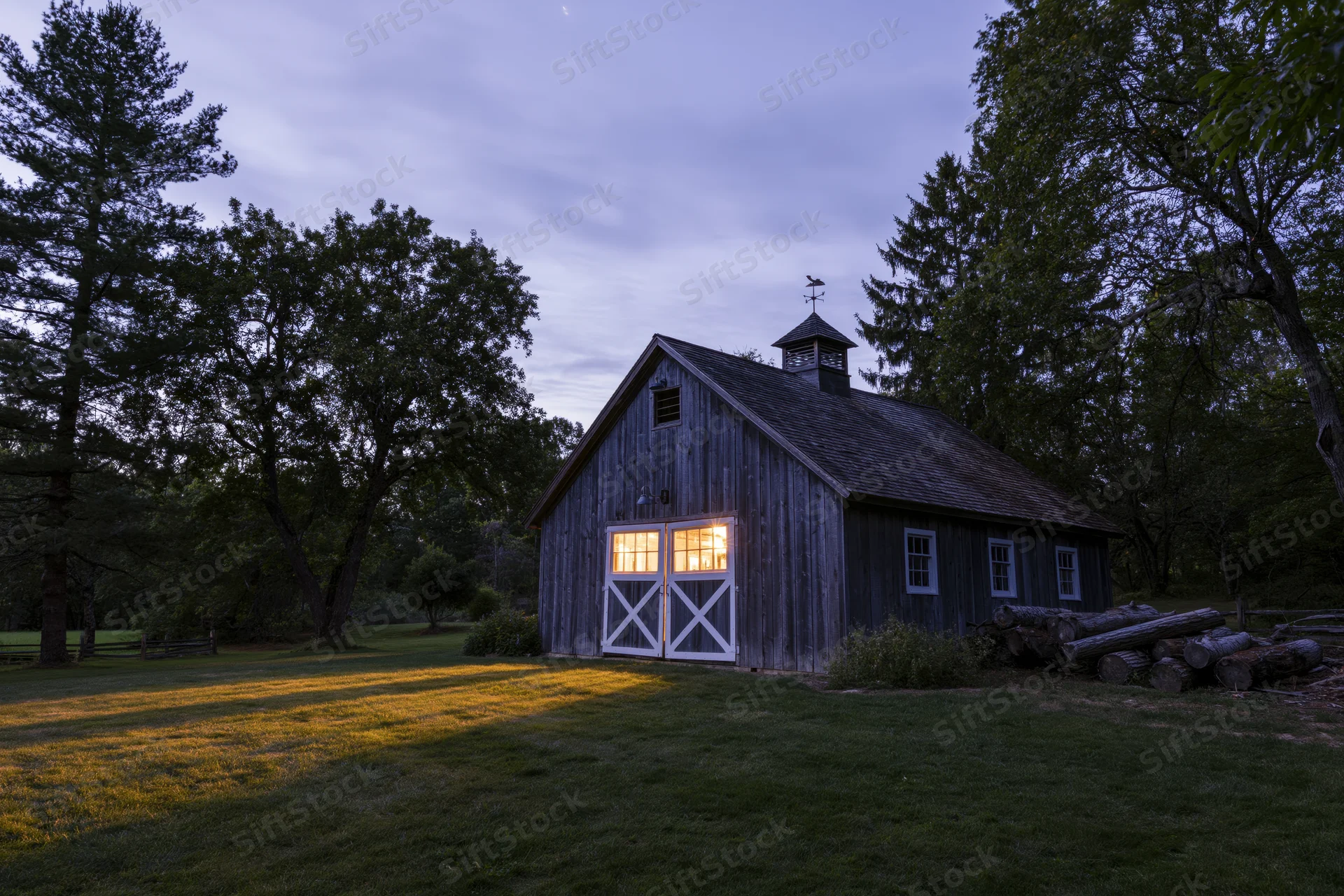 Weathered gray wooden barn with white-trimmed cross doors glowing at twilight in a grassy rural setting surrounded by trees