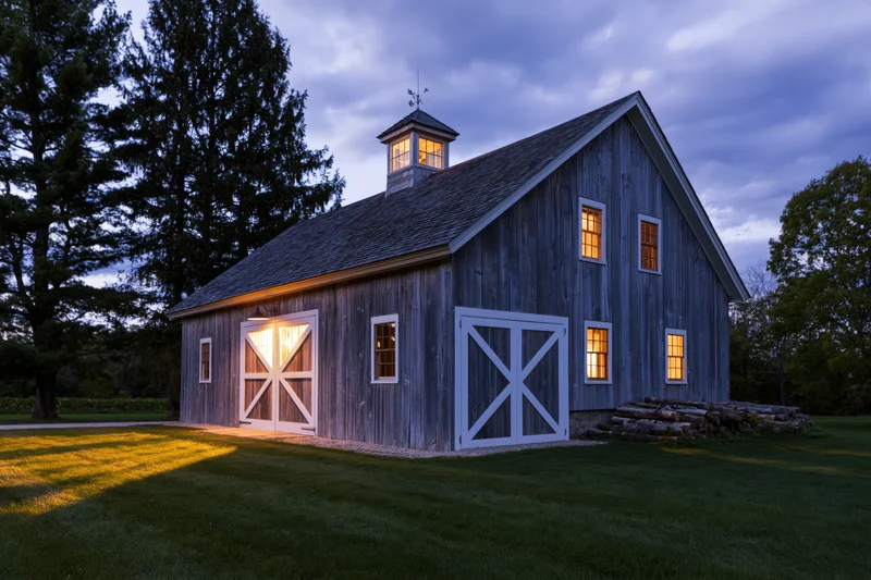 Weathered grey wooden barn with white-trimmed cross doors and glowing windows at twilight on a grassy lawn