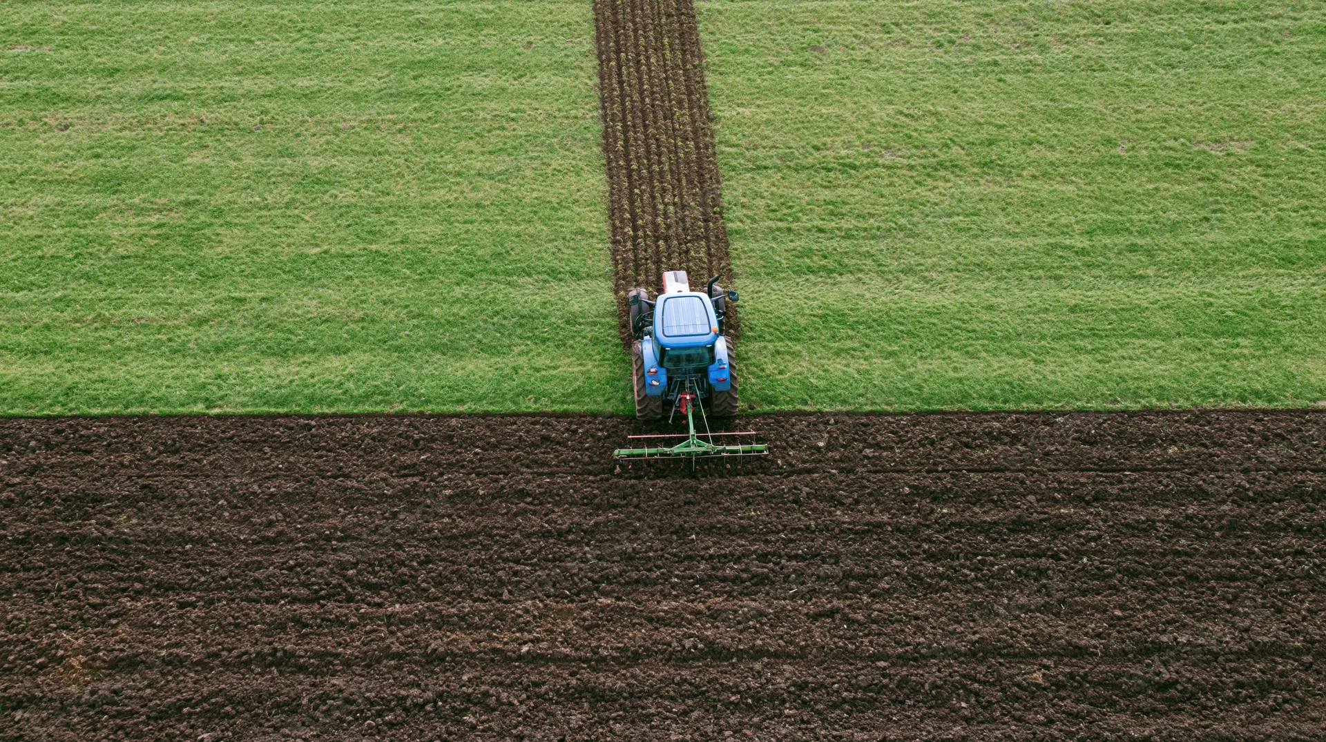 Blue tractor plowing a field from above, creating neat furrows in dark soil surrounded by green grass