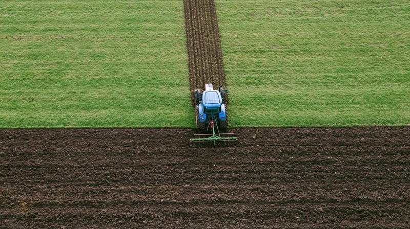 Blue tractor plowing a field from above, creating neat furrows in dark soil surrounded by green grass