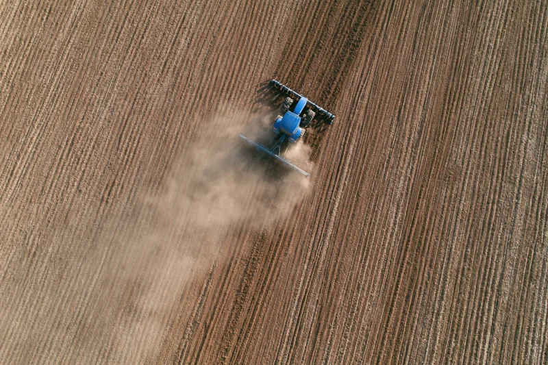 Blue tractor plowing a field from above, creating furrows and dust in a rural agricultural landscape