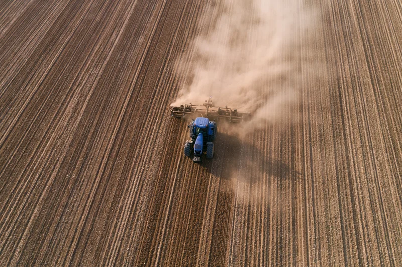 Blue tractor pulling plow through tilled field from aerial view