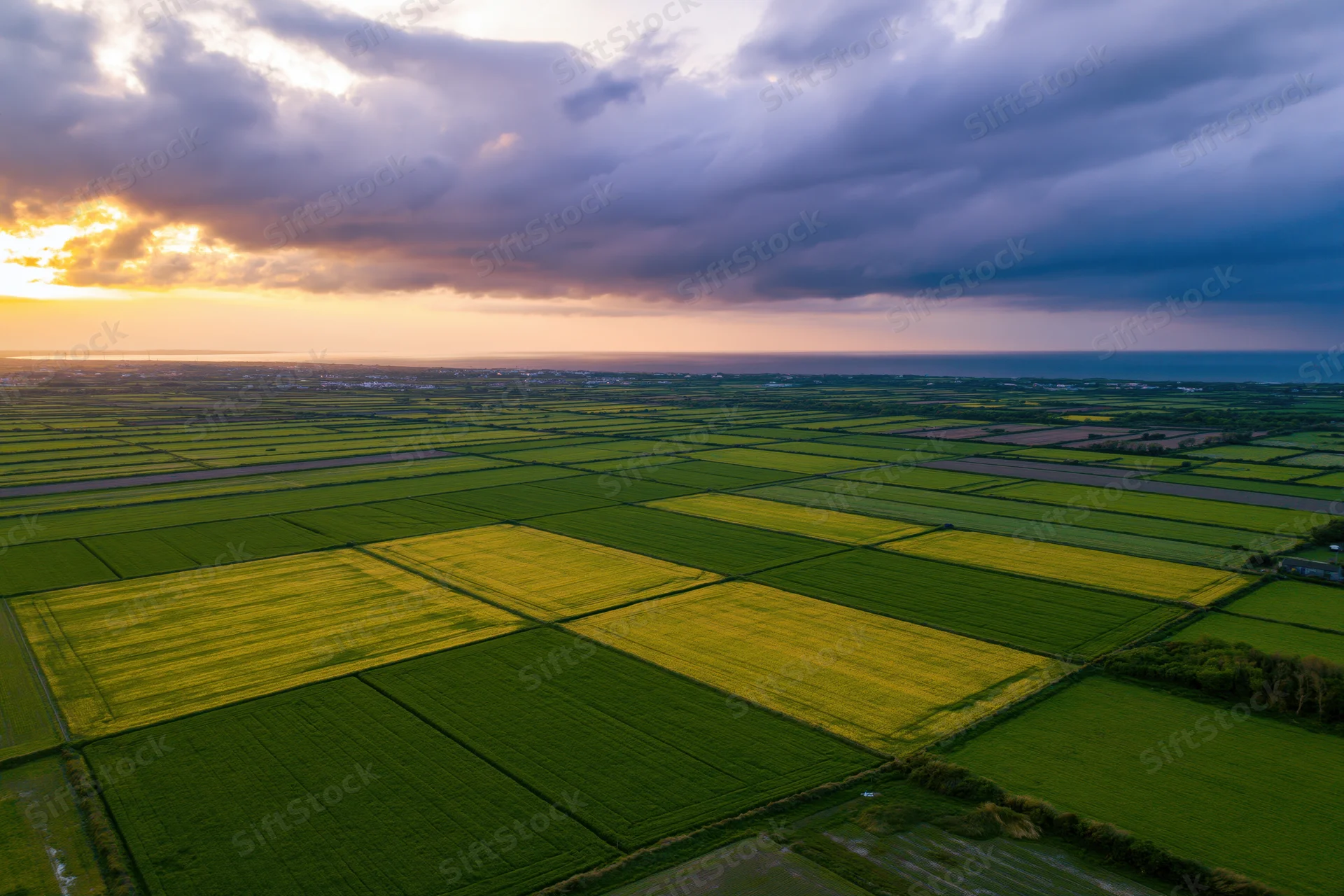 Aerial view of flat agricultural fields divided into geometric patterns under a dramatic sky at sunset near the ocean
