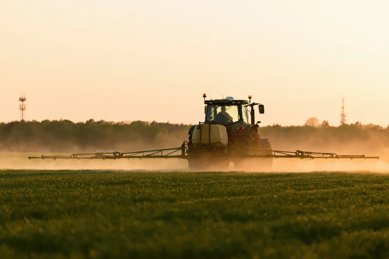 Tractor spraying pesticide across a green field at sunset