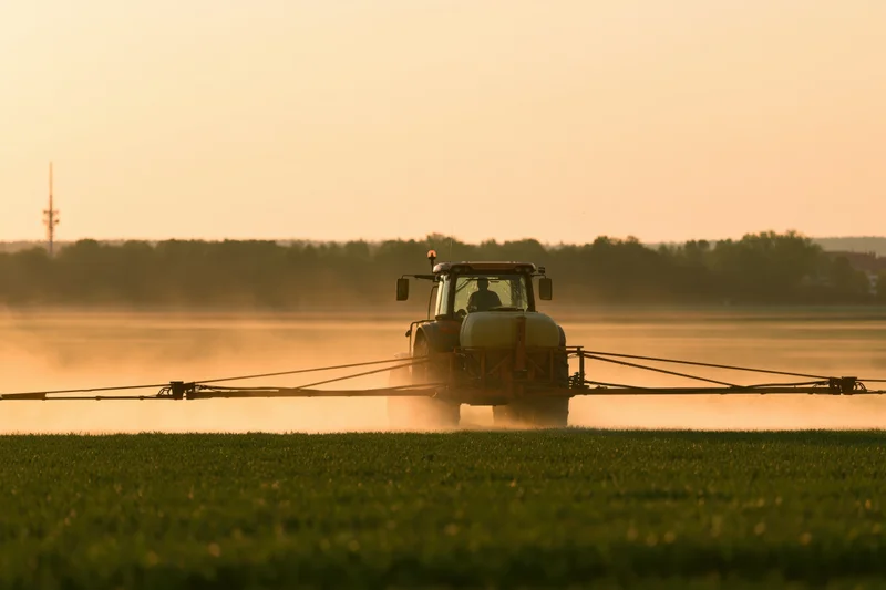 Tractor with wide spray boom applying pesticide across a green field at sunset