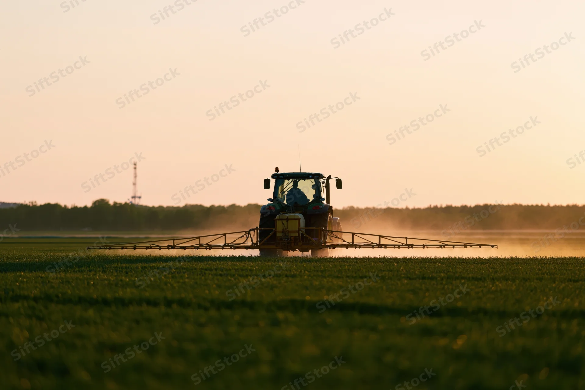 Tractor with wide spray boom applying pesticide across a green field at sunset
