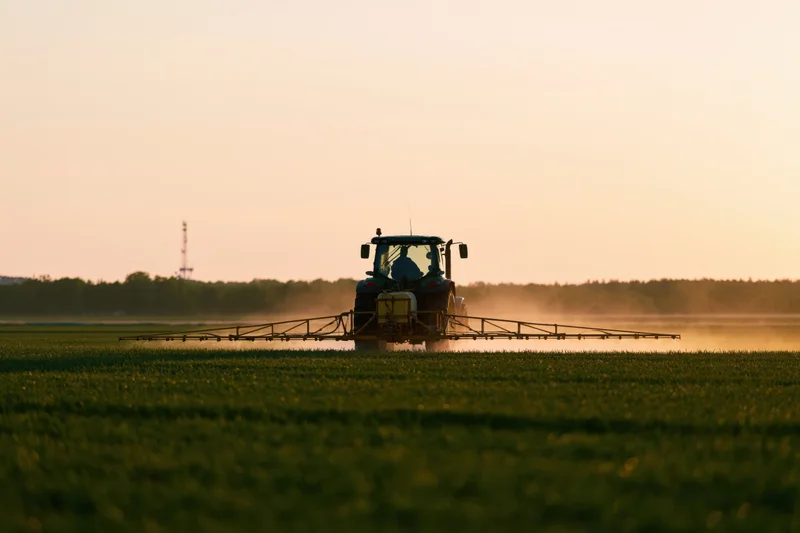 Tractor with wide spray boom applying pesticide across a green field at sunset