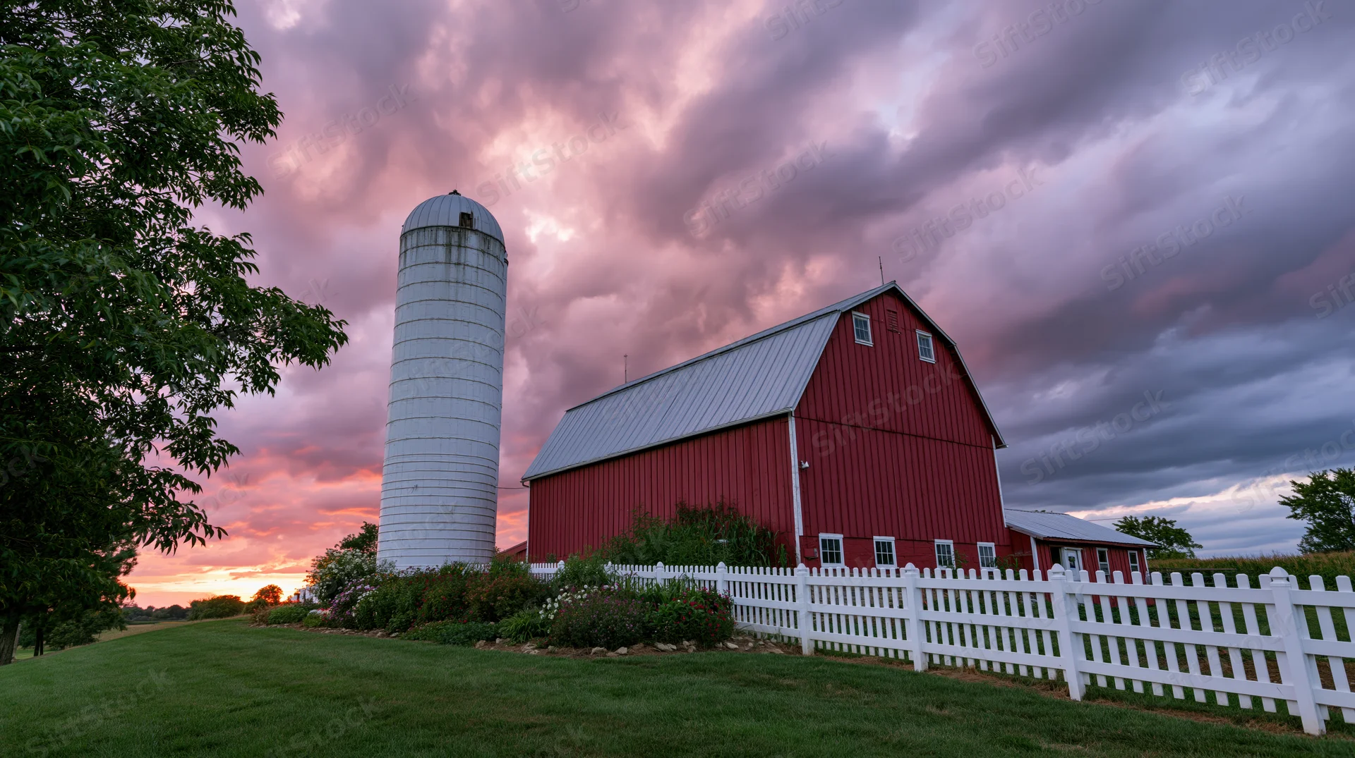 Red barn with white silo and picket fence under dramatic twilight sky