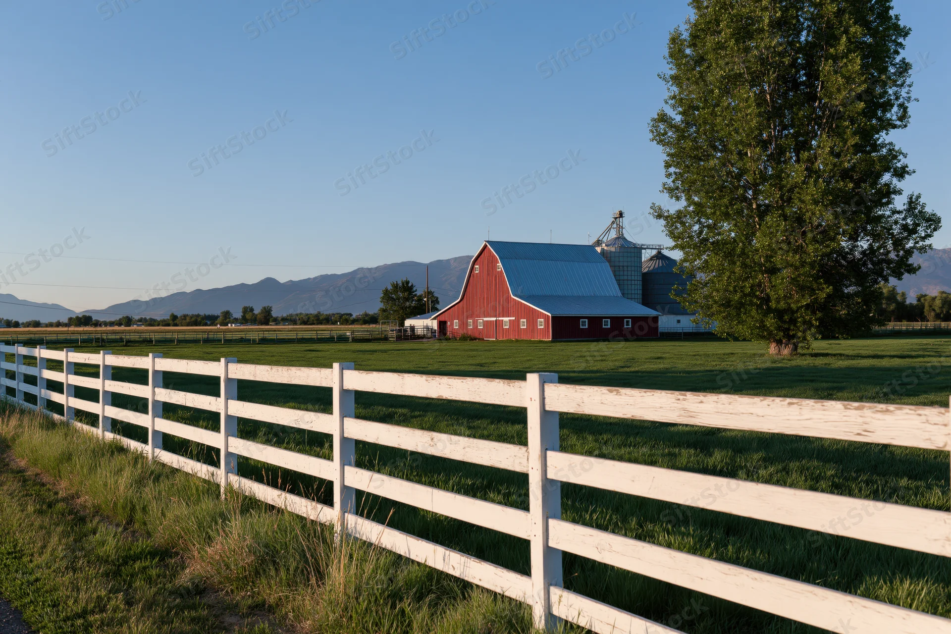 Red barn with blue metal roof in open field behind white wooden fence, surrounded by green pasture and distant mountains under clear blue sky