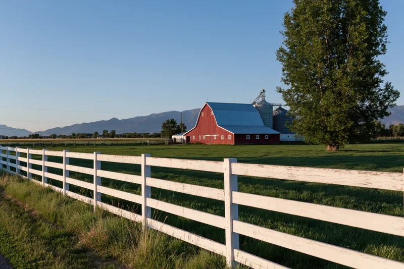 Red barn with blue metal roof in open field behind white wooden fence, surrounded by green pasture and distant mountains under clear blue sky
