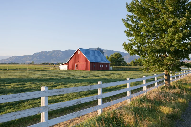 Red barn with blue metal roof in a grassy field behind a white fence with mountains in the background