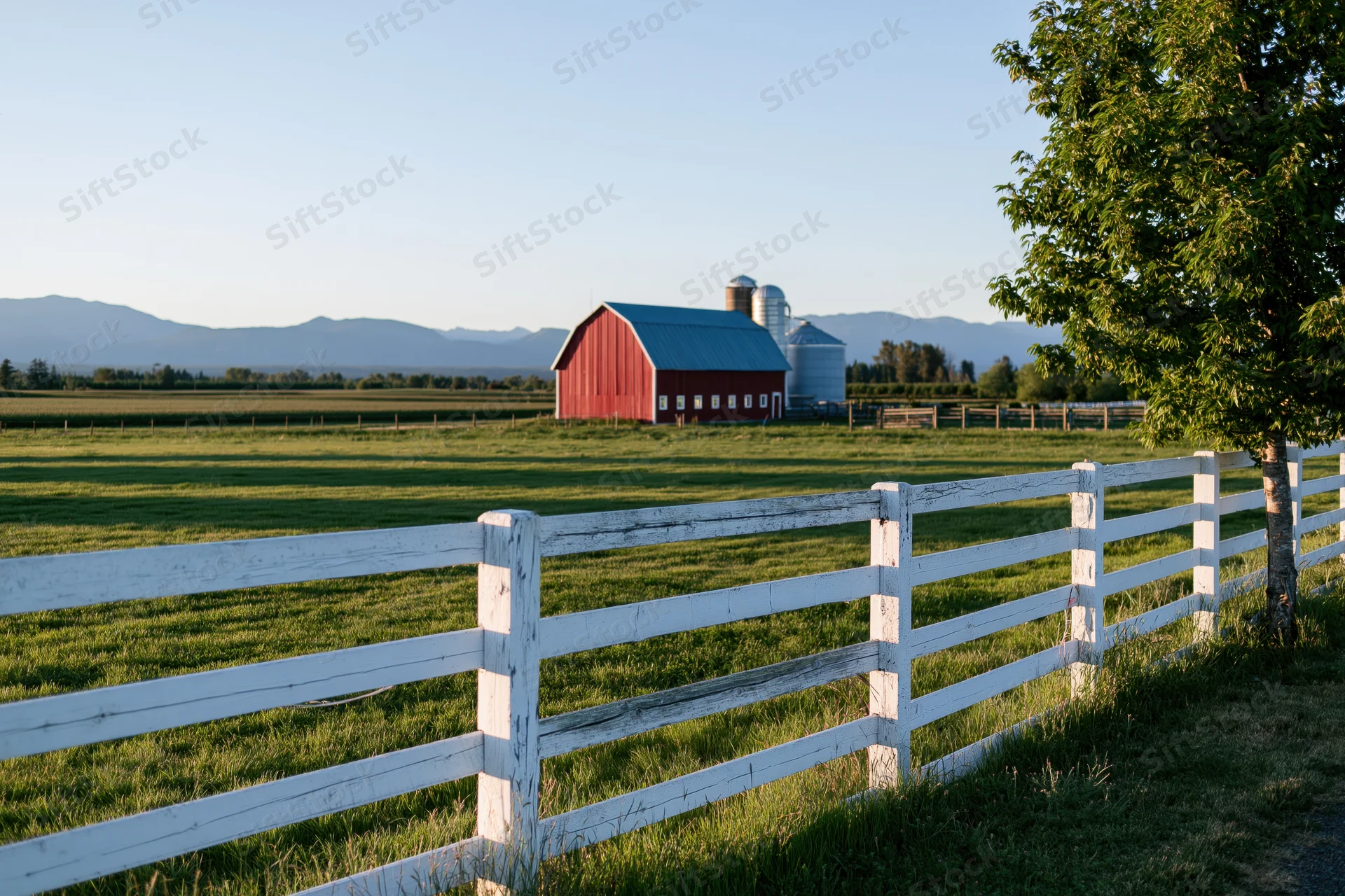 Red barn with blue metal roof and white fence in open grassy field under clear sky