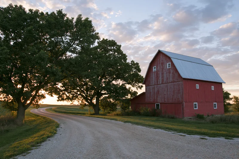 Red gambrel-roofed barn beside a winding gravel road in a rural midwestern field at sunset