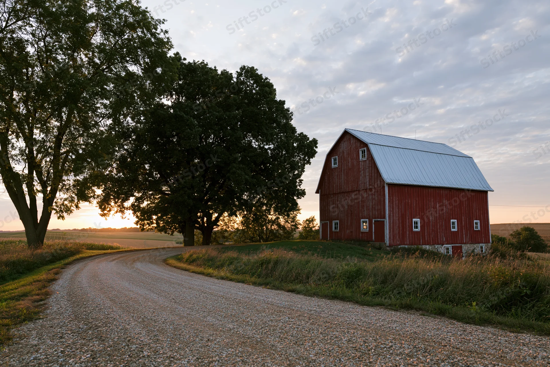Red gambrel-roofed barn on a midwestern farm at sunset beside a gravel road