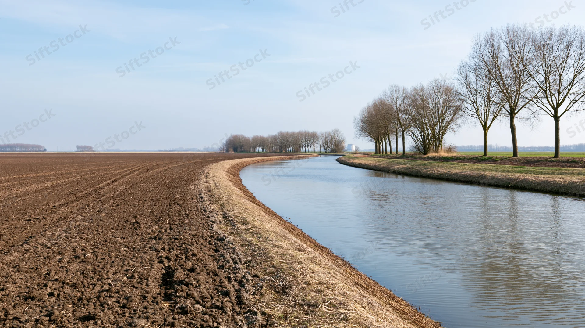 Ploughed field alongside a calm canal in a flat dutch polder landscape with bare trees under a clear sky