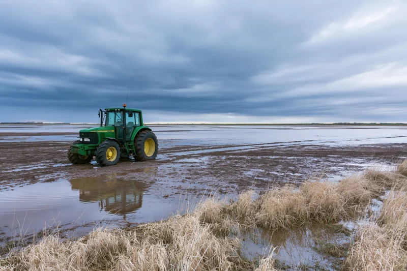 Green john deere tractor sitting idle on a waterlogged flat field under cloudy sky
