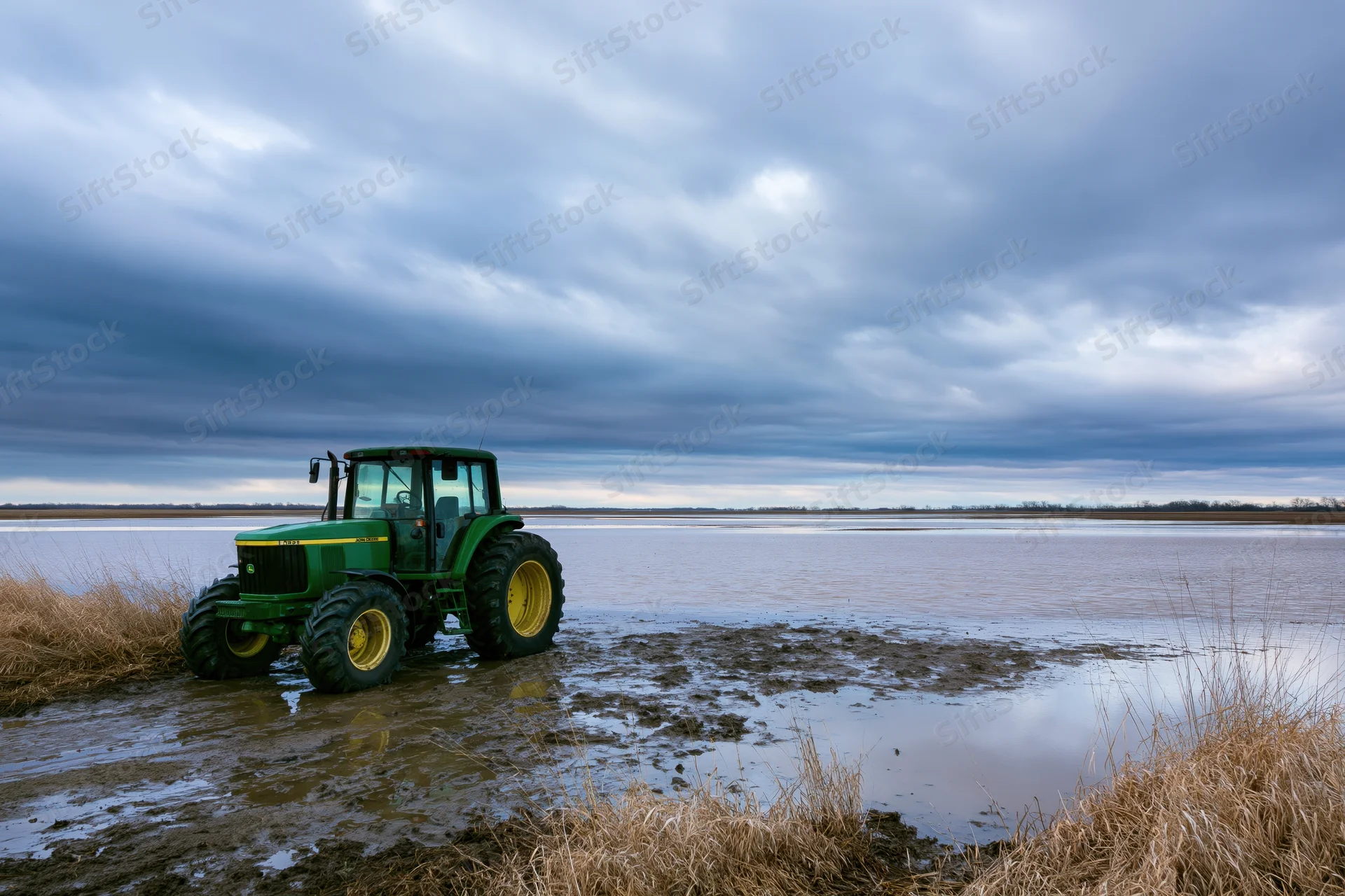 Green john deere tractor parked on muddy waterlogged field under cloudy sky