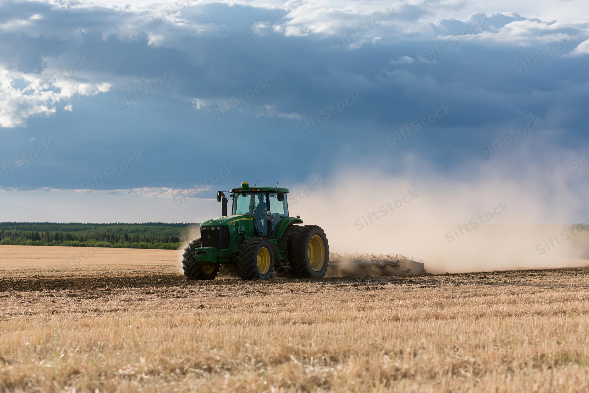 Green tractor pulling disc harrow across dry harvested field under cloudy sky