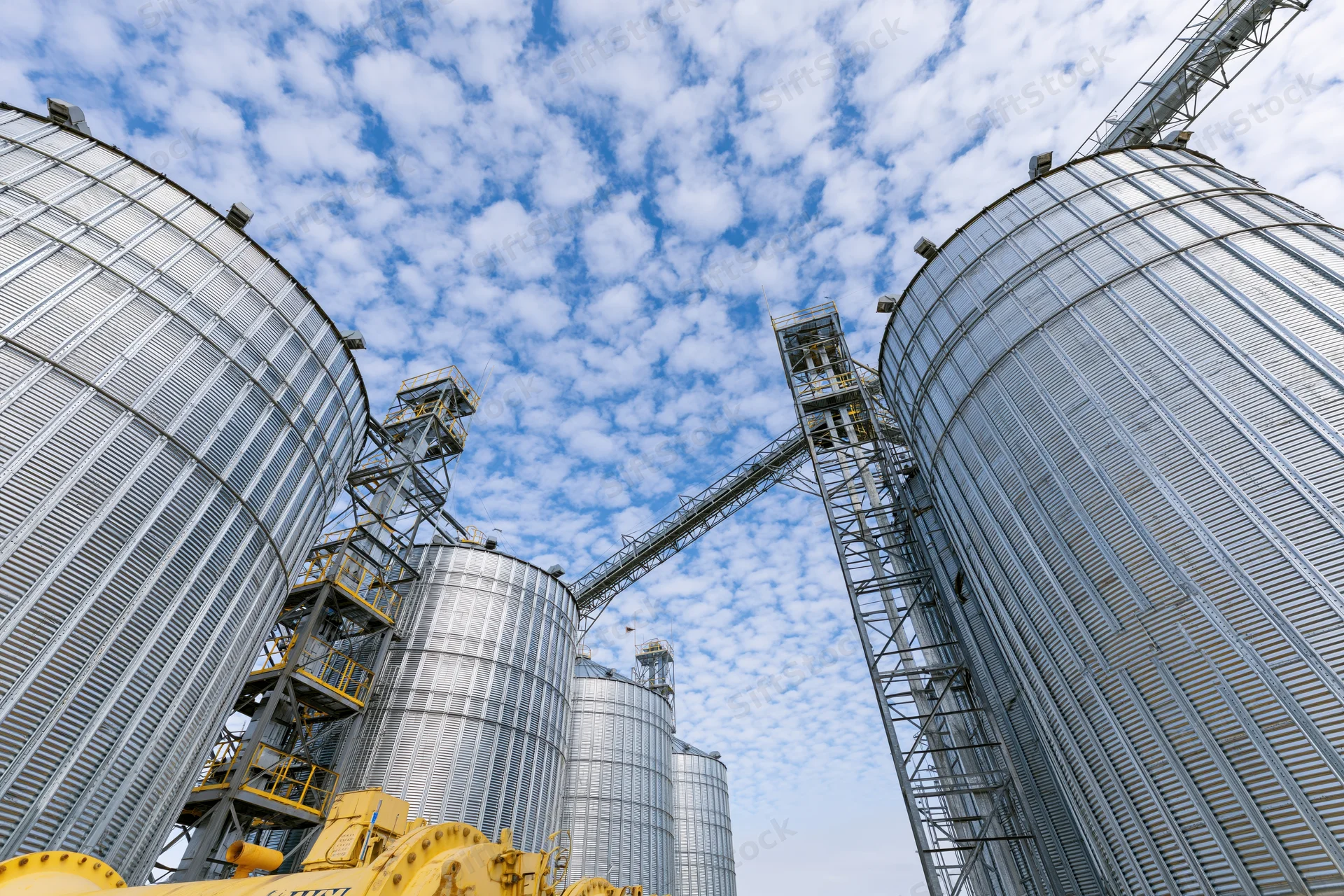 Large metal grain silos with conveyor system under partly cloudy sky