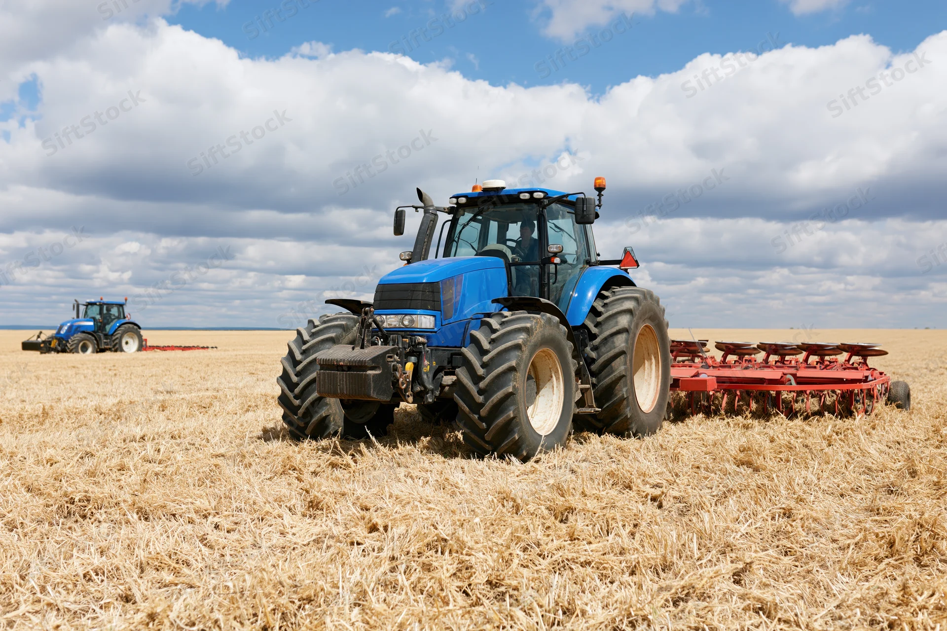 Blue tractor pulling red disc harrow across harvested field