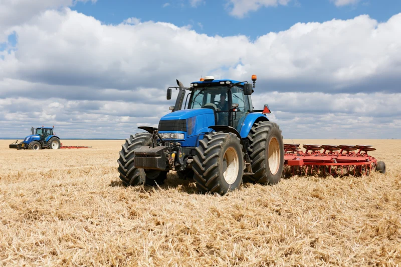 Blue tractor pulling red disc harrow across harvested field