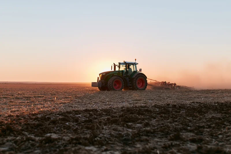 Green tractor pulling tillage equipment across a plowed field at sunset
