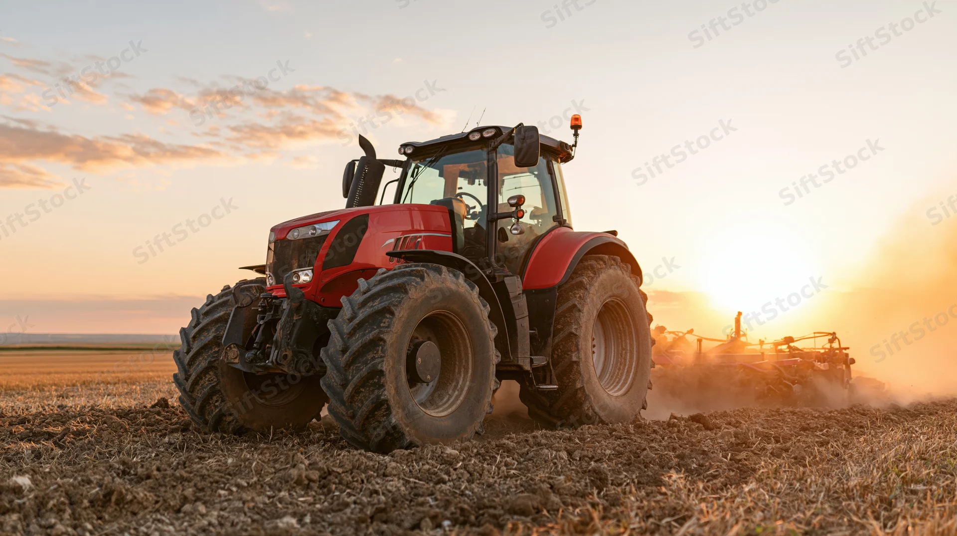 Red agricultural tractor pulling tillage implement across tilled field at sunset