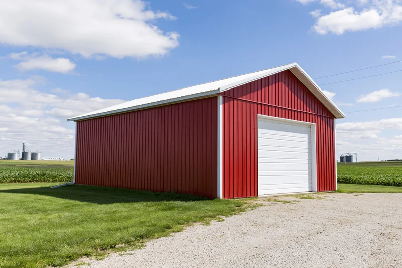 Red metal barn with white roof standing on gravel driveway beside green cornfield under blue sky with clouds