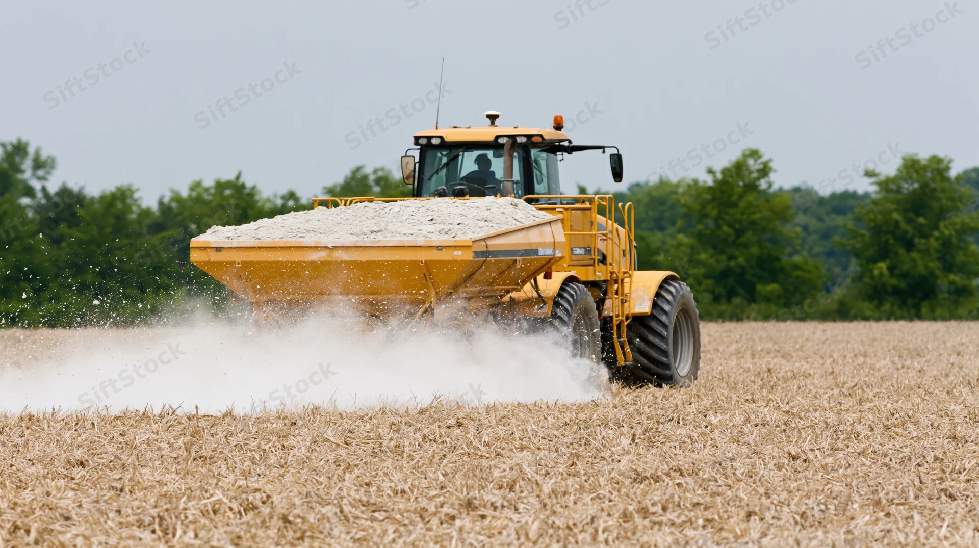 Yellow self-propelled lime spreader moving through a harvested field, dispersing lime onto soil