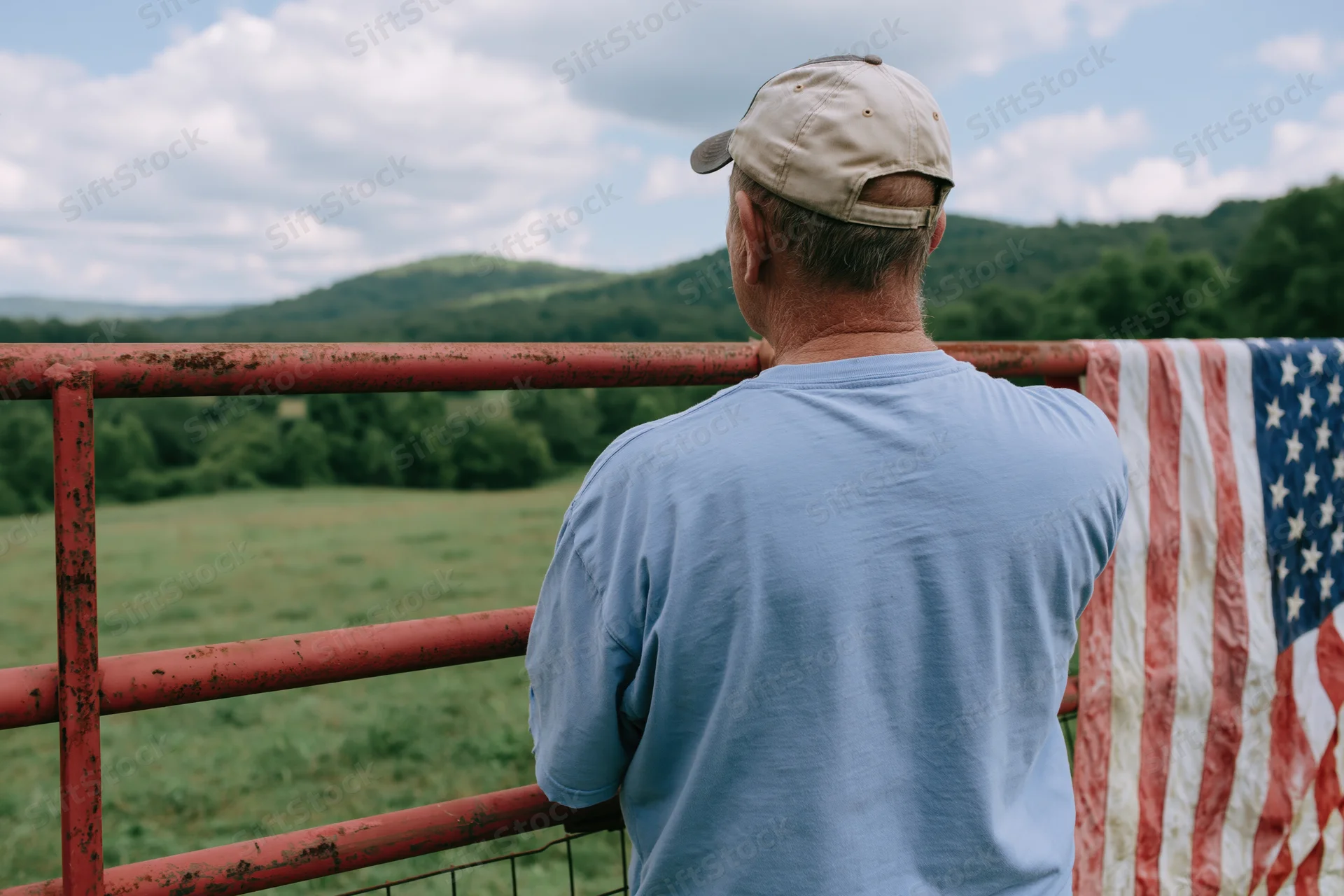 Man in baseball cap and light blue shirt standing at rusted fence overlooking green hills and fields with american flag draped beside him