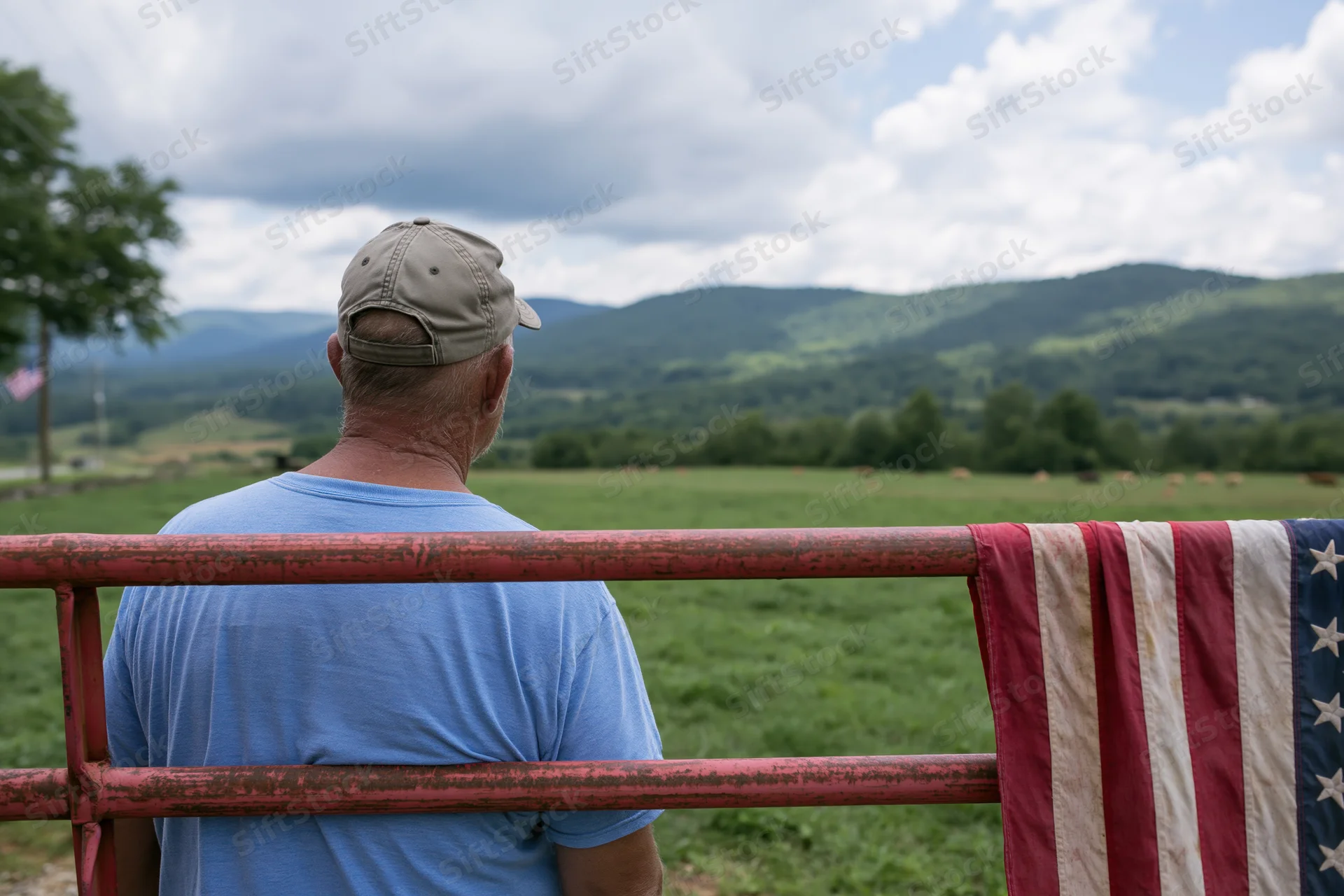 Man in baseball cap and blue tshirt standing at rusted fence overlooking green valley with american flag draped beside him