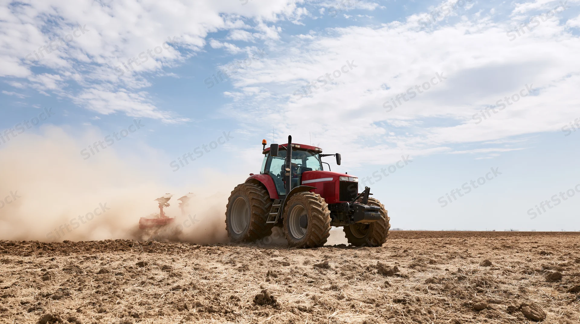 Red tractor plowing a dry tilled field under a partly cloudy sky