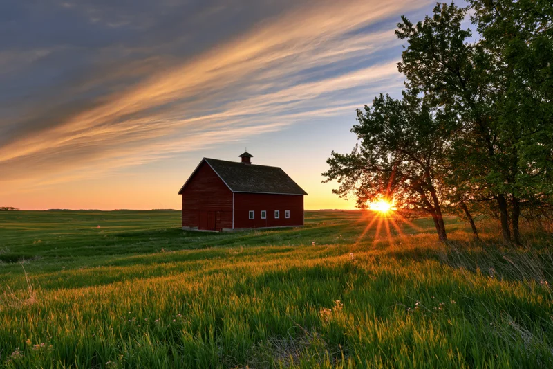 Red barn in a wide open field at sunset with trees and golden sky