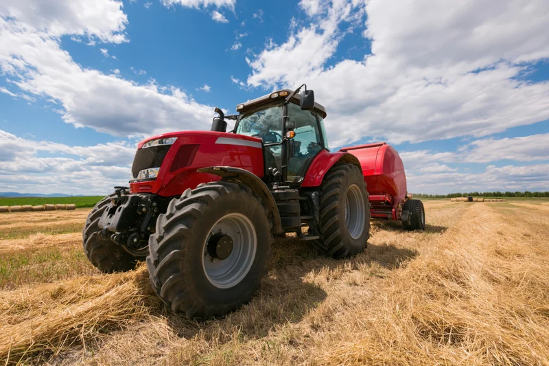Red tractor pulling round hay baler across cut hayfield under blue sky with clouds
