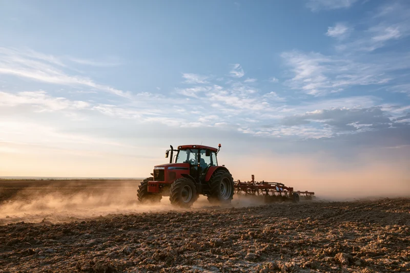 Red tractor pulling disc harrow across tilled field at sunset
