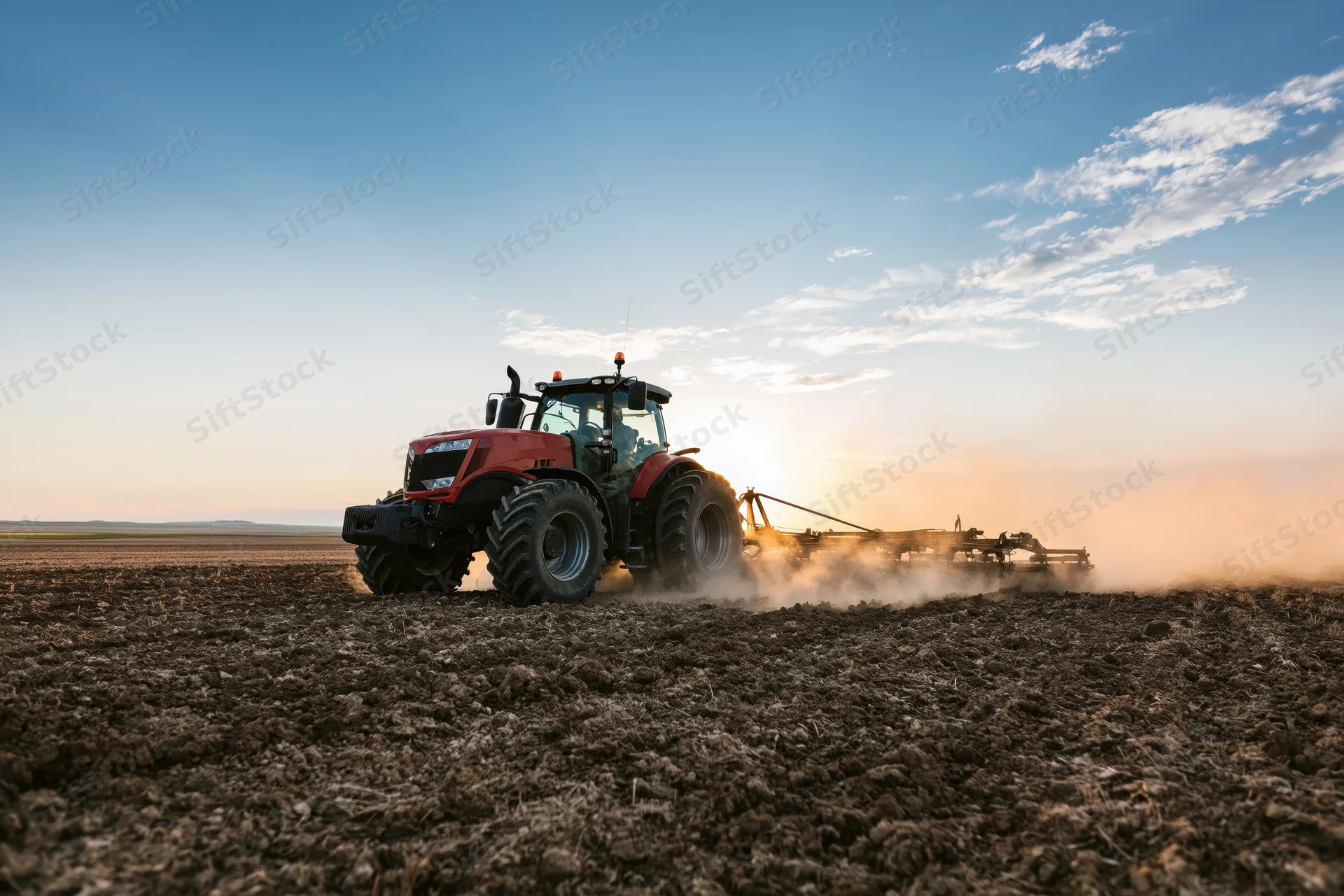Red tractor pulling disc harrow across tilled field at sunset