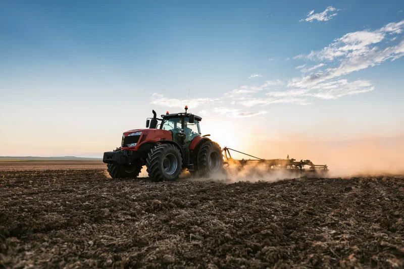 Red tractor pulling disc harrow across tilled field at sunset
