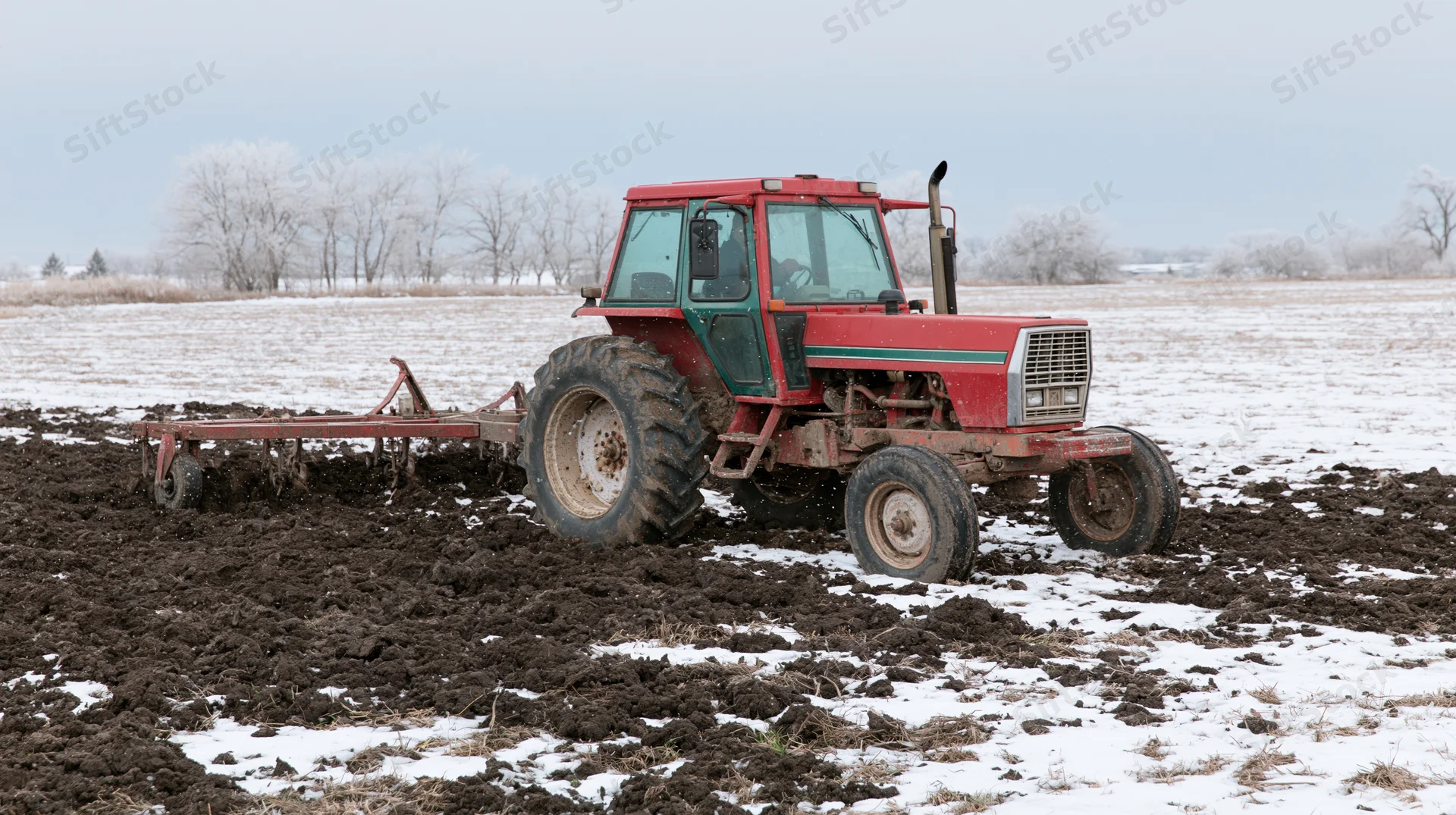 Red tractor pulling tillage equipment across a snow-dusted field in winter