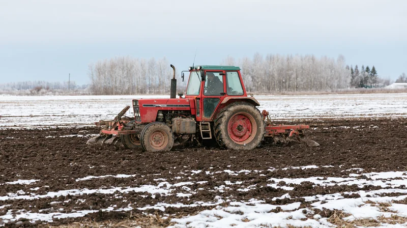 Red tractor pulling tillage equipment across a partially snow-covered field in winter