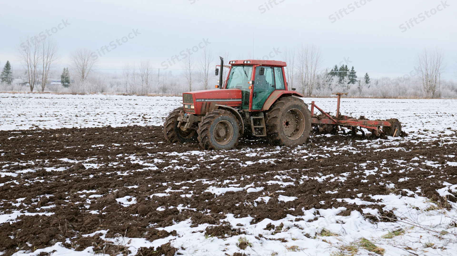 Red tractor pulling tillage equipment across snow-covered field in winter