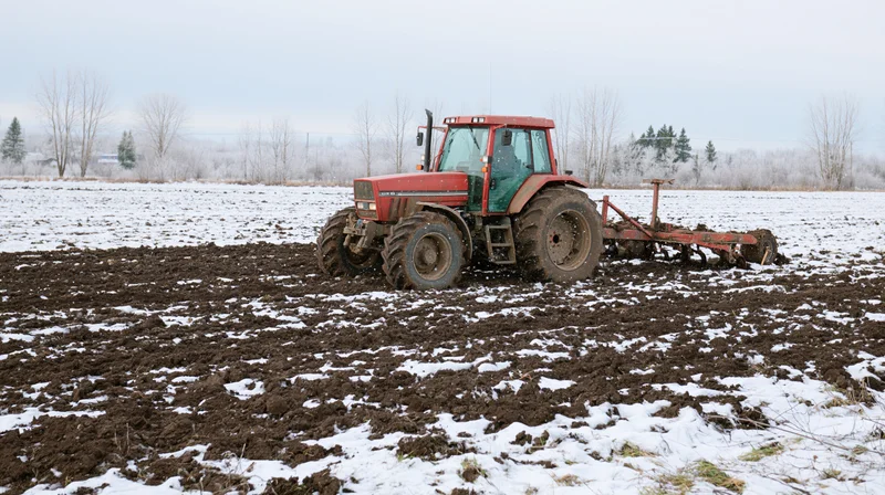 Red tractor pulling tillage equipment across snow-covered field in winter