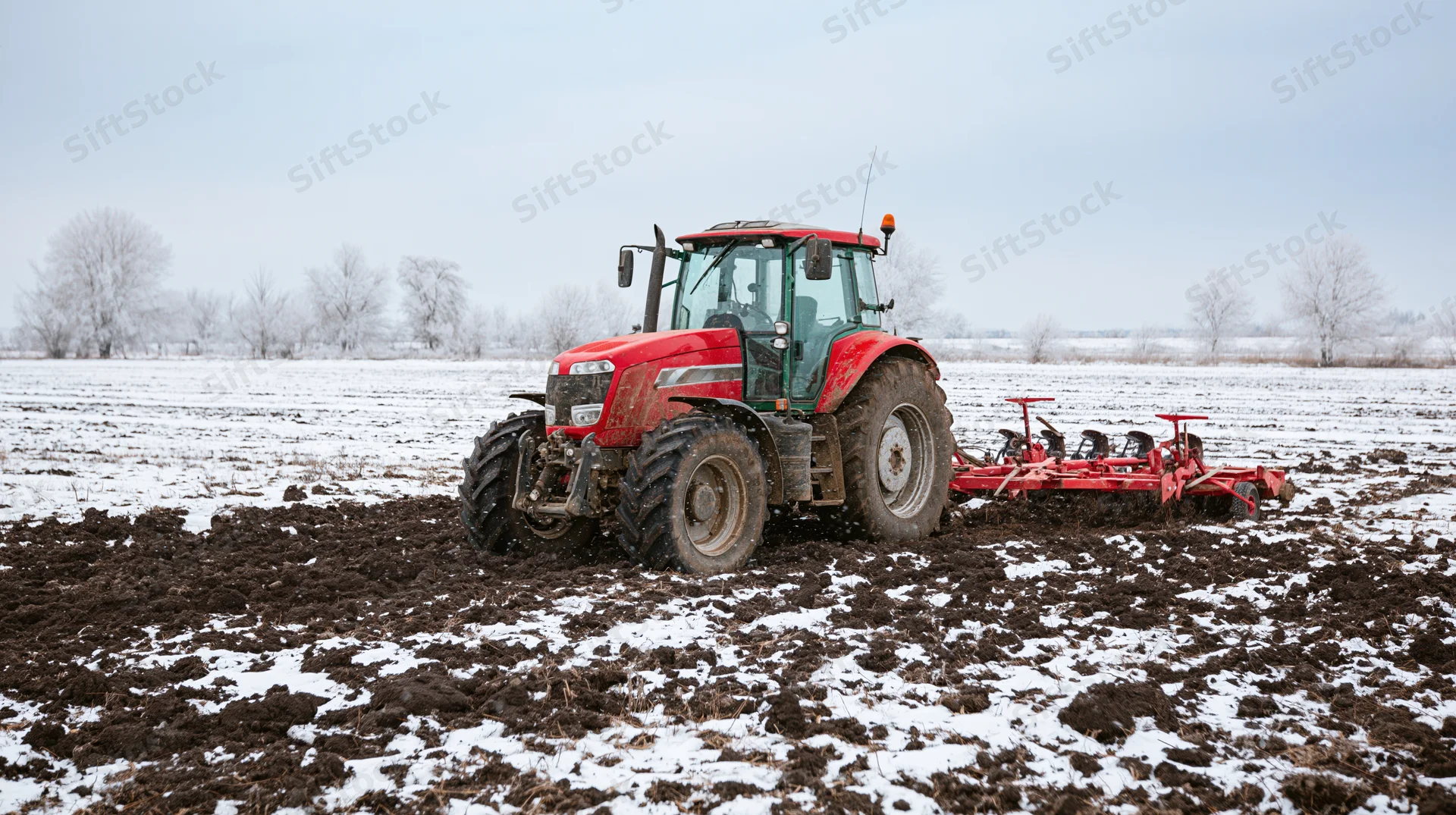 Red tractor pulling tillage equipment across a snow-covered field in winter