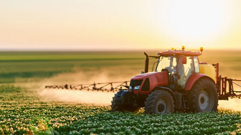 Red tractor spraying pesticide across a green crop field at sunset