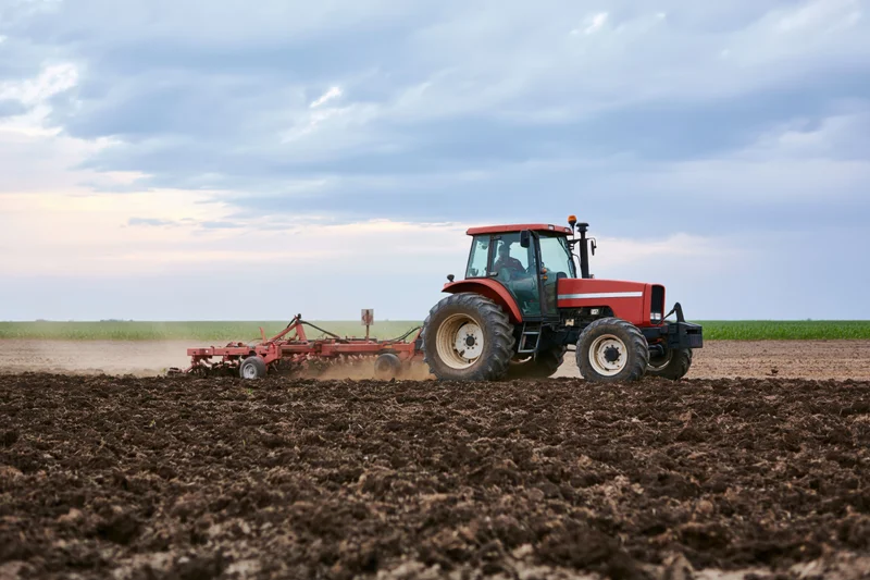 Red tractor pulling cultivator across tilled field under cloudy sky
