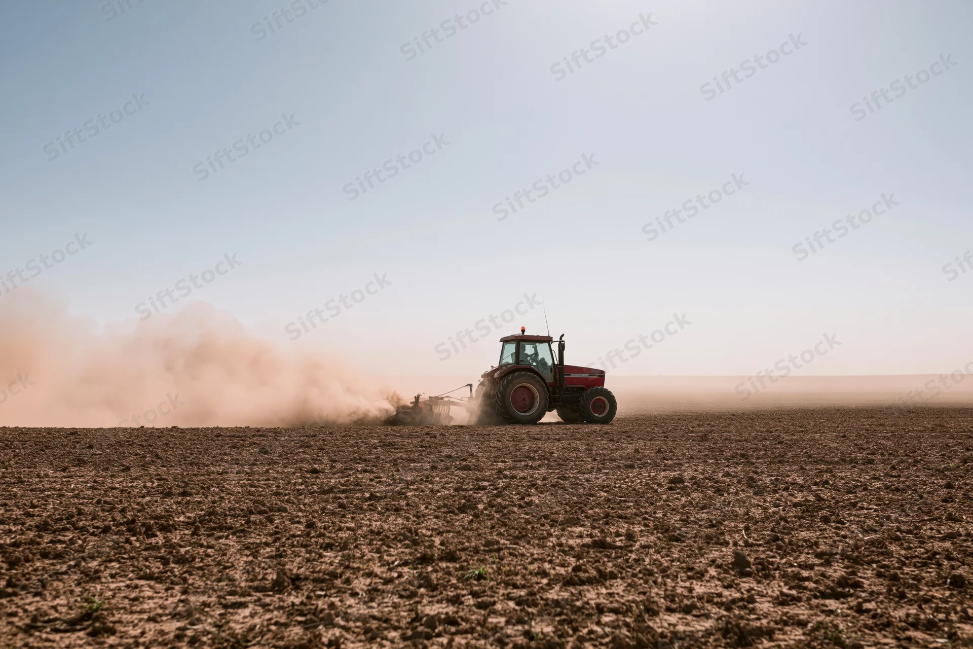 Red tractor pulling a tiller across a dry farm field kicking up dust