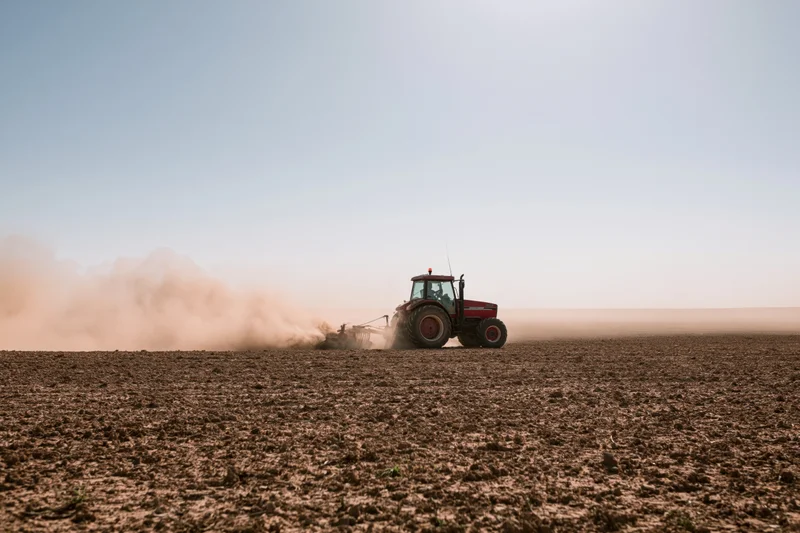 Red tractor pulling a tiller across a dry farm field kicking up dust