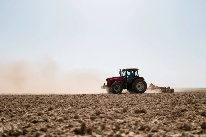 Red tractor pulling a tiller across a dry farm field kicking up dust