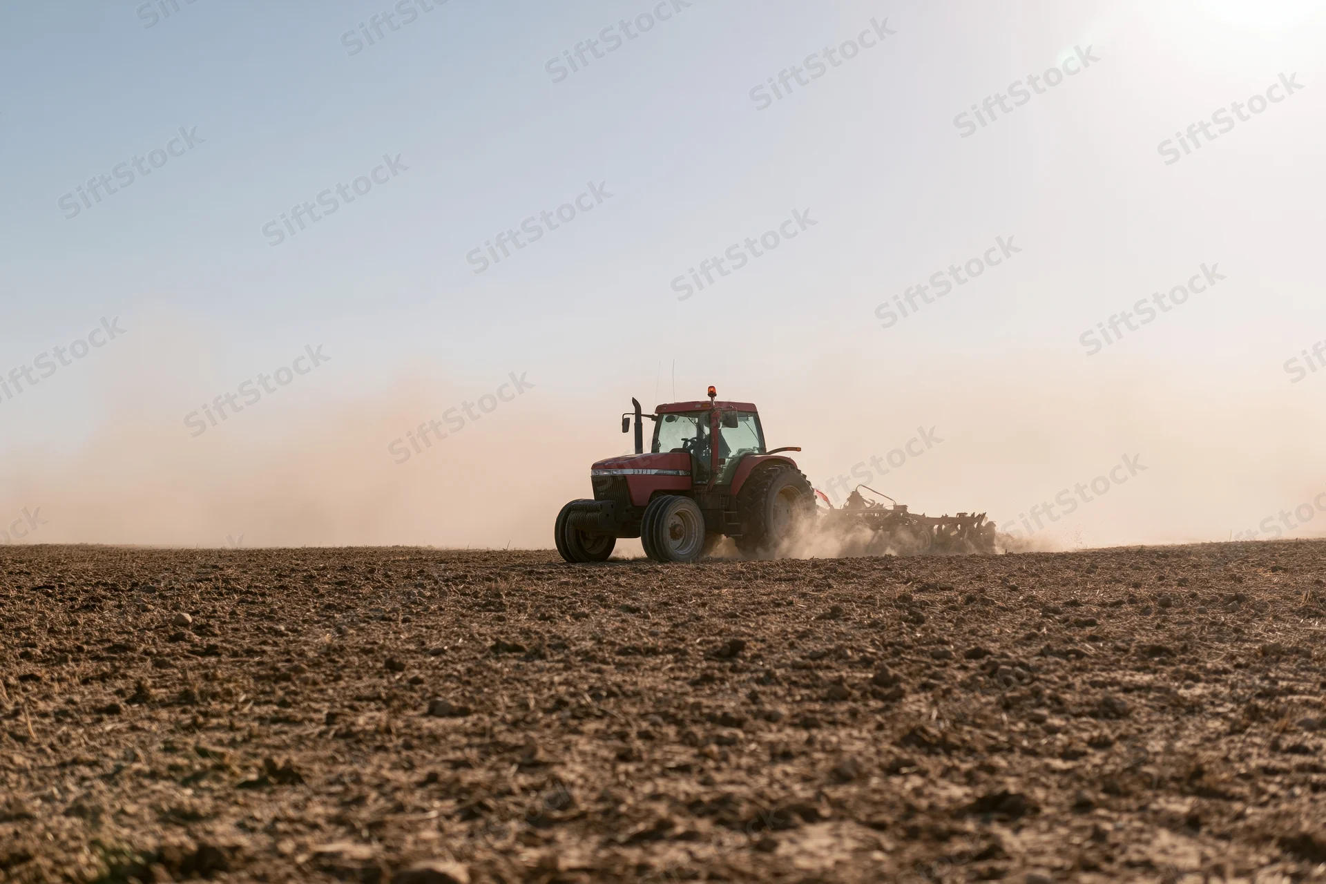 Red tractor pulling a tiller across a dry farm field kicking up dust