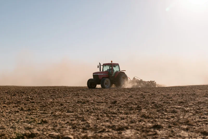 Red tractor pulling a tiller across a dry farm field kicking up dust